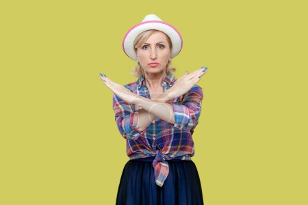 Photo for Portrait of serious senior woman wearing checkered shirt and hat crosses hands makes stop forbid sign rejects something and says no. Indoor studio shot isolated on yellow background. - Royalty Free Image