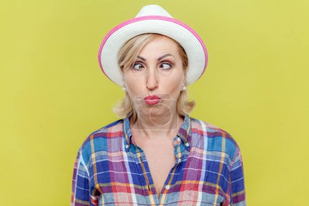 Portrait of funny foolish mature woman wearing checkered shirt and hat making grimace and crosses eyes, playing fool, having fun. Indoor studio shot isolated on yellow background.