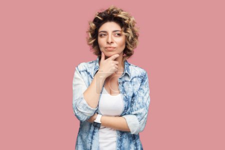 Photo for Portrait of puzzled thoughtful woman with curly hairstyle wearing blue shirt standing holding chin, thinking, planing something. Indoor studio shot isolated on pink background. - Royalty Free Image