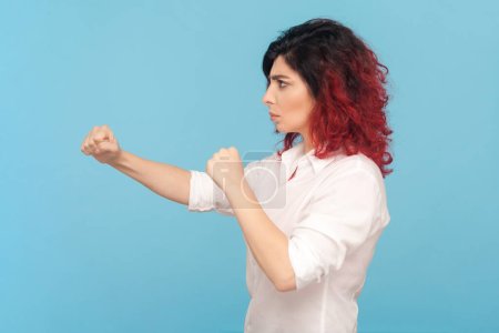 Photo for Side view portrait of strict arguing woman with fancy red hair standing with clenched fists, boxing, fighting, wearing white shirt. Indoor studio shot isolated on blue background. - Royalty Free Image