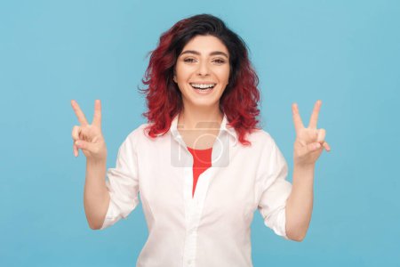 Photo for Portrait of joyful cheerful happy smiling woman with fancy red hair showing peace gesture, demonstrating v sign, wearing white shirt. Indoor studio shot isolated on blue background. - Royalty Free Image