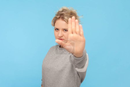 Photo for Portrait of strict serious blonde woman raised her arm showing stop gesture forbidden sign looking at camera, wearing gray sweatshirt. Indoor studio shot isolated on blue background. - Royalty Free Image