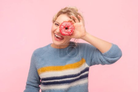 Photo for Portrait of funny playful positive blonde woman wearing sweatshirt covering her eye with tasty delicious donut smiling with tongue out. Indoor studio shot isolated on light pink background. - Royalty Free Image