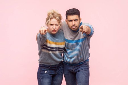 Portrait of angry irritated woman and man standing pointing together at camera arguingg with somebody expressing hate. Indoor studio shot isolated on light pink background.