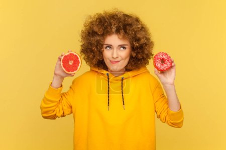 Photo for Portrait of hungry crazy woman with Afro hairstyle holding tasty doughnut and half of grapefruit, showing tongue out, wearing casual style hoodie. Indoor studio shot isolated on yellow background. - Royalty Free Image