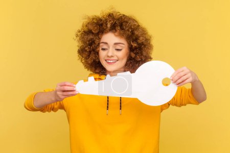 Photo for Portrait of delighted cheerful woman with Afro hairstyle holding in hand big paper key as symbol of her own apartment, state housing program. Indoor studio shot isolated on yellow background. - Royalty Free Image