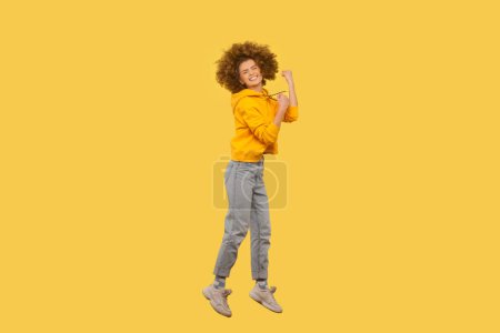 Téléchargez les photos : Oui, je l'ai fait. Portrait complet d'une femme ravie avec une coiffure afro sautant dans les airs avec des poings serrés, portant un sweat à capuche de style décontracté. Studio intérieur tourné isolé sur fond jaune. - en image libre de droit