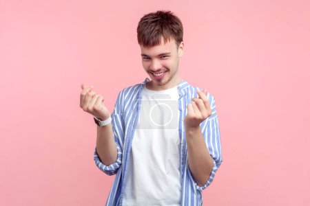 Portrait of smiling sly satisfied young man wearing shirt standing showing money gesture asking his reward salary. Indoor studio shot isolated on pink background.