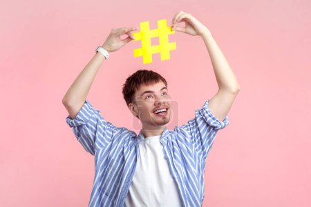 Foto de Retrato de feliz joven guapo sonriente con camisa que sostiene el hashtag amarillo por encima de la cabeza mirando hacia fuera las tendencias de Internet. Estudio interior plano aislado sobre fondo rosa. - Imagen libre de derechos