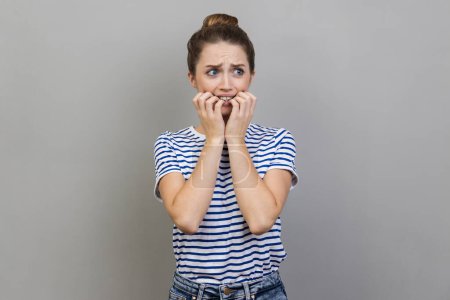 Photo for Portrait of woman wearing striped T-shirt biting nails, being nervous terrified, feeling frightened of challenge to start business, anxiety disorder. Indoor studio shot isolated on gray background. - Royalty Free Image