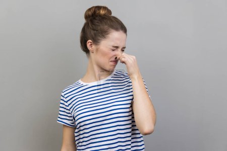 Téléchargez les photos : Répulsion à l'odeur puante. Portrait de femme portant un T-shirt rayé retenant son souffle, se pincant le nez et grimaçant de dégoût, évitant les mauvaises odeurs. Studio intérieur tourné isolé sur fond gris. - en image libre de droit
