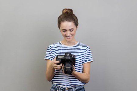 Photo for Portrait of satisfied delighted woman traveler or photographer wearing striped T-shirt holding and looking at photocamera, taking photo enjoying hobby. Indoor studio shot isolated on gray background. - Royalty Free Image