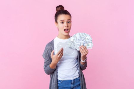Photo for Portrait of confused teenage girl with bun hairstyle in casual clothes standing pointing big sum of money holding dollar banknotes. Indoor studio shot isolated on pink background. - Royalty Free Image
