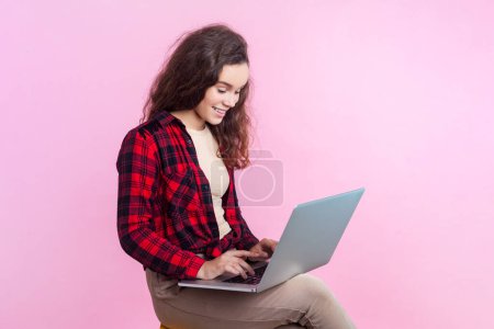 Téléchargez les photos : Portrait d'adolescente souriante pigiste aux cheveux ondulés en chemise à carreaux rouge assise tapant sur un clavier d'ordinateur portable écrit article. Studio intérieur tourné isolé sur fond rose. - en image libre de droit