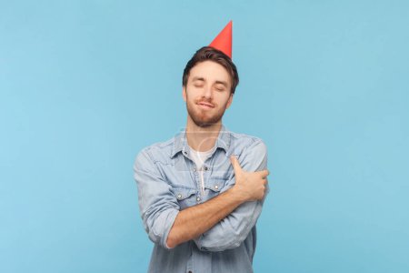 Photo for Portrait of happy satisfied unshaven birthday man in party cone celebrating holiday, hugging himself, egoist, wearing denim shirt. Indoor shot isolated on blue background. - Royalty Free Image