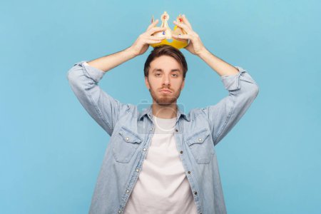 Photo for Portrait of satisfied delighted man with bristle putting on golden crown, looking with arrogance and smile, privileged status, wearing denim shirt. Indoor shot isolated on blue background. - Royalty Free Image