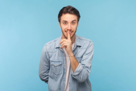 Photo for Portrait of smiling mysterious unshaven man presses index finger to lips makes hush gesture tells secret asks to be quiet, wearing denim shirt. Indoor shot isolated on blue background. - Royalty Free Image