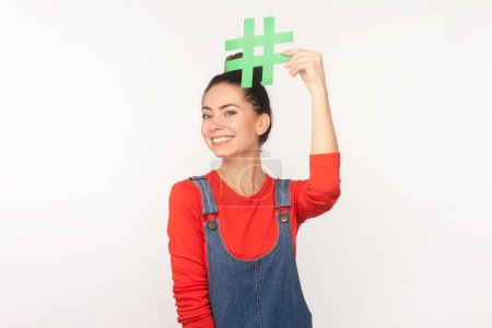 Photo for Portrait of woman with hair bun holding hashtag symbol above head, promoting viral topic in social network, tagging blog trends, wearing denim overalls. Indoor studio shot isolated on white background - Royalty Free Image