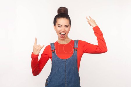 Photo for Portrait of woman with hair bun makes rock n roll gesture shows she is cool feels very happy, screaming enjoyable, wearing denim overalls. Indoor studio shot isolated on white background - Royalty Free Image