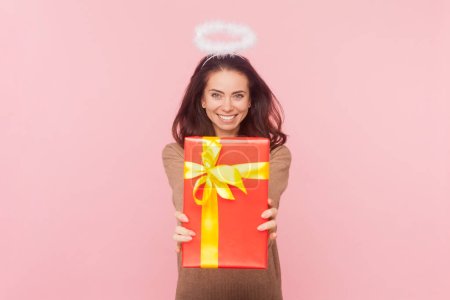 Portrait de joyeuse femme heureuse aux cheveux ondulés et nimbe au-dessus de la tête donnant boîte cadeau enveloppé rouge, félicitant, portant pull marron. Studio intérieur tourné isolé sur fond rose