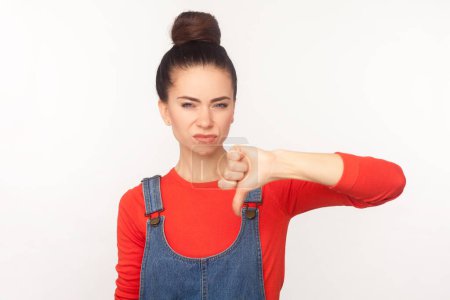 Photo for Portrait of sad disappointed woman with hair bun frowning face showing thumb down dislike gesture disapprove sign, wearing denim overalls. Indoor studio shot isolated on white background - Royalty Free Image