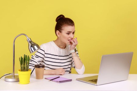 Photo for Nervous woman office worker biting her nails sitting at workplace and looking at laptop screen with worry expression, scared of software error. Indoor studio studio shot isolated on yellow background. - Royalty Free Image