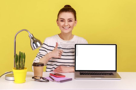 Photo for Smiling happy young woman employee pointing empty laptop screen, mock up display for advertisement, showing thumb up, website promotion. Indoor studio studio shot isolated on yellow background. - Royalty Free Image