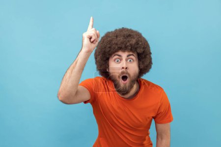 Téléchargez les photos : Portrait d'homme émerveillé avec une coiffure afro portant un T-shirt orange pointant du doigt et regardant émerveillé par une idée de génie soudaine, une solution obtenue, prise de vue en studio intérieure isolée sur fond bleu. - en image libre de droit