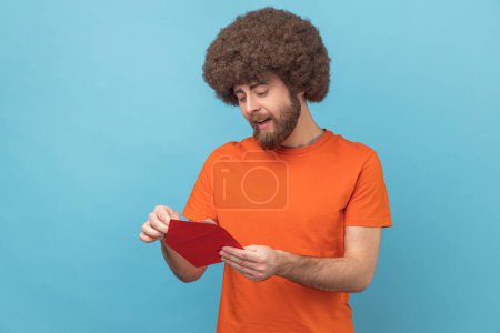 Téléchargez les photos : Portrait d'homme à la coiffure afro portant un T-shirt orange lisant une lettre ou une carte de vœux, tenant une enveloppe, souriant et réjouissant d'agréables nouvelles. Studio intérieur tourné isolé sur fond bleu. - en image libre de droit