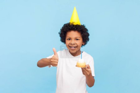 Téléchargez les photos : Portrait d'un petit garçon d'anniversaire aux cheveux bouclés portant un cône jaune tenant un délicieux beignet savoureux avec une bougie montrant le pouce levé comme un geste. Studio intérieur tourné isolé sur fond bleu. - en image libre de droit