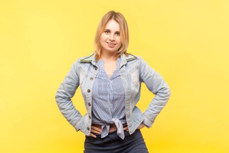 Photo for Portrait of good looking charming blonde woman wearing denim jacket standing with hands on hips looking at camera with satisfied expression. Indoor studio shot isolated on yellow background. - Royalty Free Image