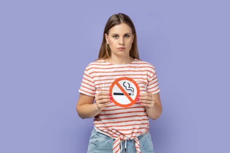 Photo for Portrait of serious beautiful woman wearing striped T-shirt holding no smoking sign and looking at camera with strict face, take care of your health. Indoor studio shot isolated on purple background. - Royalty Free Image