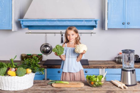 Téléchargez les photos : Enfant blonde tenant brocoli et chou-fleur souriant regardant la caméra, debout dans la cuisine moderne préparant une salade de légumes, cuisinant un plat sain, aide sa mère à dîner. - en image libre de droit
