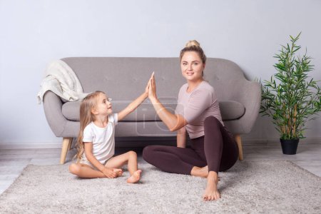 Téléchargez les photos : Portrait de belle femme confiante avec coiffure de chignon donnant haute cinq à la fille après l'entraînement ensemble, assis sur le sol après des séances de yoga, grand rôle parental et maternité. - en image libre de droit