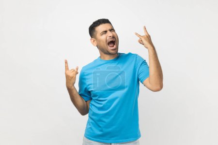 Photo for Portrait of excited amazed young adult unshaven man wearing blue T- shirt standing showing rock and roll gesture, spending time on festival. Indoor studio shot isolated on gray background. - Royalty Free Image