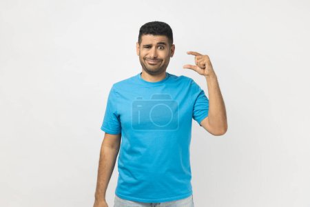 Photo for Portrait of confused unshaven man wearing blue T- shirt standing looking at camera with frowning face, showing small size with fingers. Indoor studio shot isolated on gray background. - Royalty Free Image