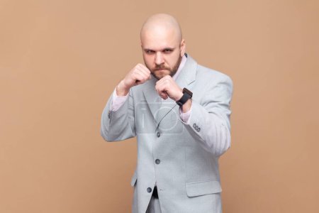 Boxing and self-defence. Portrait of angry aggressive bald bearded man keeping fists clenched, ready for fight, wearing gray jacket. Indoor studio shot isolated on brown background.