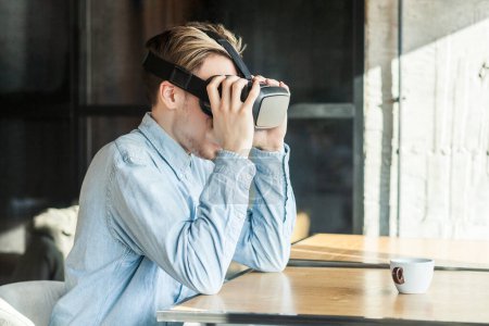 Photo for Side view portrait of young adult man in blue denim shirt sitting in cafe, wearing vr and watching video on simulator holding headset resting while having break. Indoor shot. - Royalty Free Image