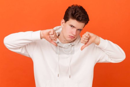 Photo for Portrait of young man wearing white hoodie expresses dislike with body language, keeps thumb down and frowns face, has displeased face. Indoor studio shot isolated on orange background. - Royalty Free Image