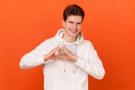 Photo for Portrait of pleased good looking man wearing white hoodie shapes heart over chest, confesses in love, has romantic mood, expresses truthful feelings. Indoor studio shot isolated on orange background. - Royalty Free Image