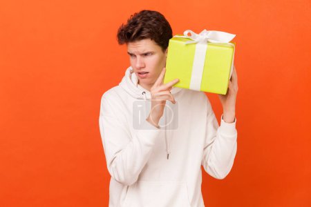 Photo for Portrait of curious brunette handsome young man wearing white hoodie holding shaking present box, trying to guess what inside. Indoor studio shot isolated on orange background. - Royalty Free Image