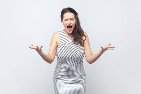 Photo for Portrait of annoyed outraged attractive brunette woman gestures angrily and shouts loudly expresses rage and hate, wearing striped dress. Indoor studio shot isolated on gray background. - Royalty Free Image