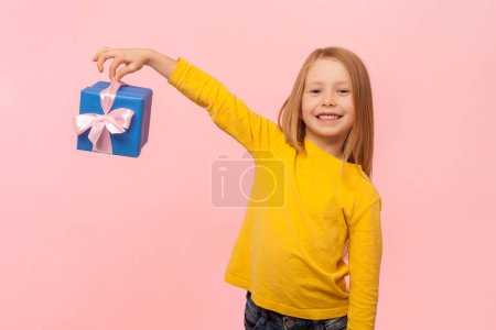 Photo for Portrait of cheerful blonde little girl standing with blue wrapped gift box, looking at camera with happy expression, wearing yellow jumper. Indoor studio shot isolated on pink background. - Royalty Free Image