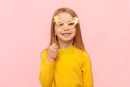 Photo for Portrait of smiling satisfied little girl smiling to camera, holding party props, paper glasses, having fun on birthday party, wearing yellow jumper. Indoor studio shot isolated on pink background. - Royalty Free Image