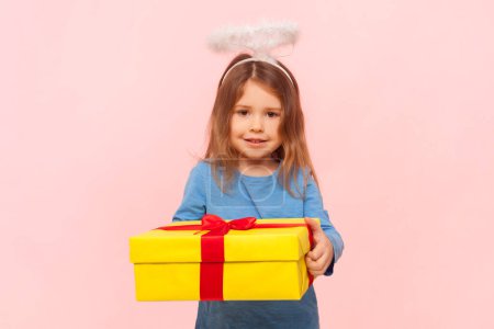 Portrait of adorable little girl with nimb over head holding big yellow present box, preparing gift for friend, wearing blue sweater. Indoor studio shot isolated on pink background.