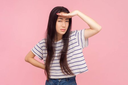 Photo for Portrait of attentive serious woman with long brunette hair standing keeps hand near forehead expecting something, wearing striped T-shirt. Indoor studio shot isolated on pink background. - Royalty Free Image