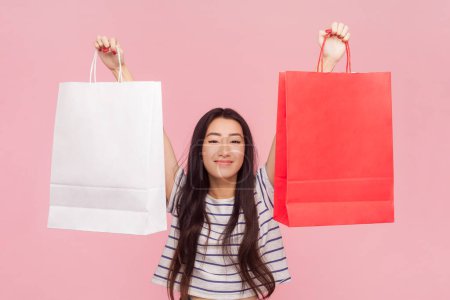 Photo for Portrait of positive beautiful woman with long brunette hair showing shopping bags visiting fashion mall, wearing striped T-shirt. Indoor studio shot isolated on pink background. - Royalty Free Image