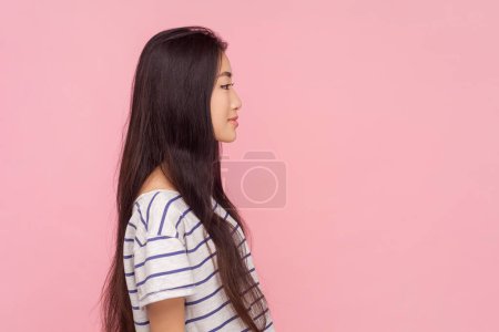 Téléchargez les photos : Portrait latéral d'une femme souriante aux longs cheveux bruns regardant vers l'avenir avec un sourire d'expression positive, portant un T-shirt rayé. Studio intérieur tourné isolé sur fond rose. - en image libre de droit