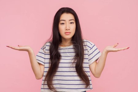 Photo for Portrait of puzzled woman with long brunette hair standing spreads hands having uncertain expression, shrugging shoulders, wearing striped T-shirt. Indoor studio shot isolated on pink background. - Royalty Free Image