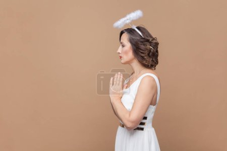 Photo for Portrait of hopeful middle aged woman with wavy hair and nimb over head, standing with palms together, praying gesture, wearing white dress. Indoor studio shot isolated on light brown background. - Royalty Free Image